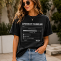 A woman wearing a faded black graphic t-shirt with a computer dialog box design that reads 'EXPORTING MY FEELINGS.WAV' and 'rendering emotions.