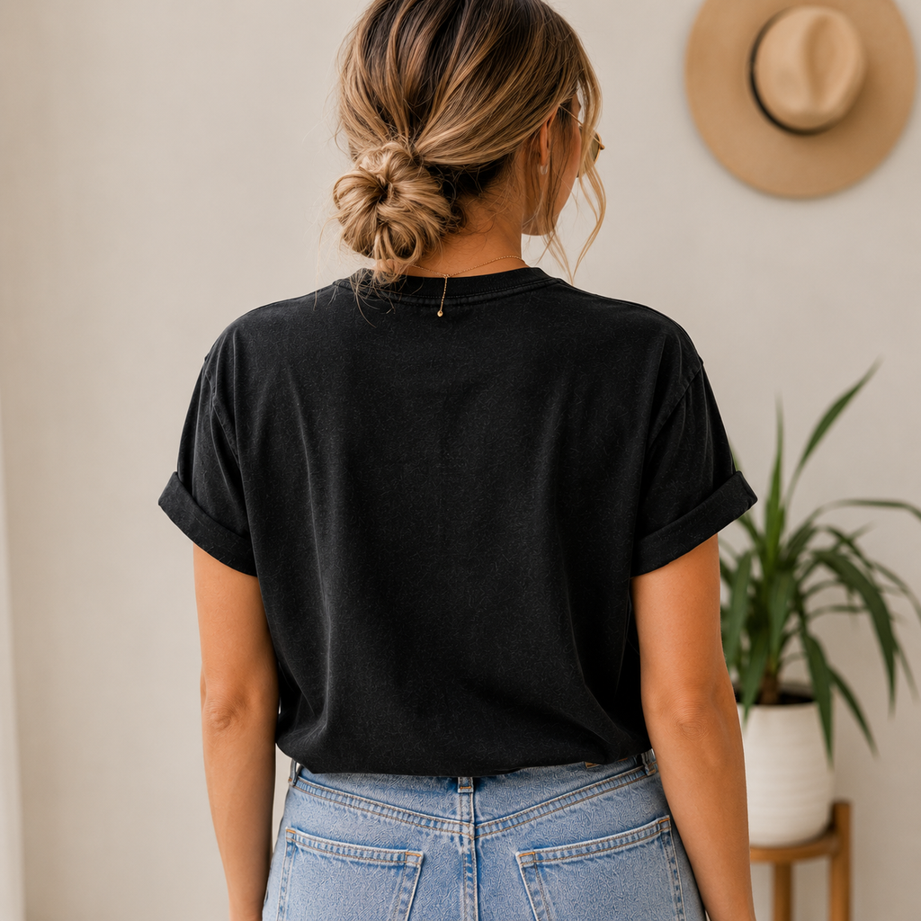 A woman viewed from behind wearing a black short-sleeve t-shirt with rolled cuffs and light-wash blue jeans, standing in a brightly lit indoor setting with a hat and potted plant in the background.
