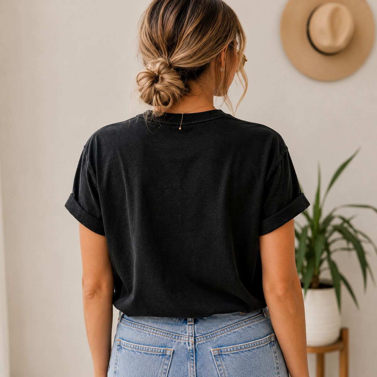 A woman viewed from behind wearing a black short-sleeve t-shirt with rolled cuffs and light-wash blue jeans, standing in a brightly lit indoor setting with a hat and potted plant in the background.