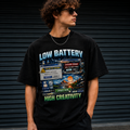 A man wearing a black graphic t-shirt with a retro computer error screen design that reads 'LOW BATTERY' and 'HIGH CREATIVITY', posed in front of a dark metal shutter.
